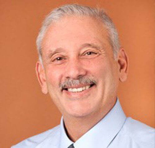 A smiling man with gray hair and mustache in a light blue shirt, set against a warm brown background.