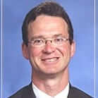 Smiling man in formal attire with glasses, blue background. Professional portrait, suitable for medical, business, or academic context.