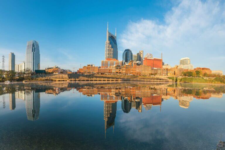 Nashville downtown skyline with iconic buildings reflecting in the Cumberland River, clear blue sky background.