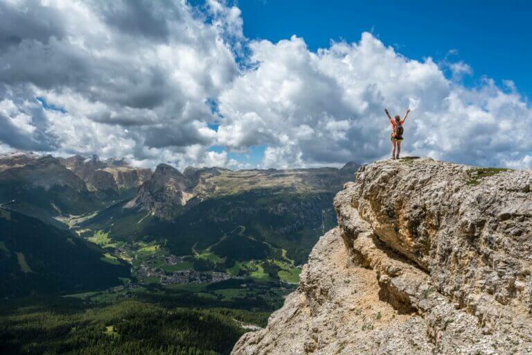 Hiker triumphantly stands on rocky cliff edge overlooking scenic mountain valley under dramatic cloudy sky.