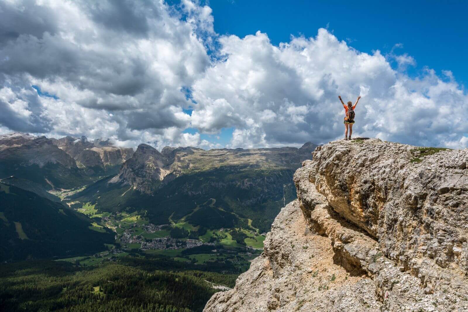 Hiker triumphantly stands on rocky cliff edge overlooking scenic mountain valley under dramatic cloudy sky.