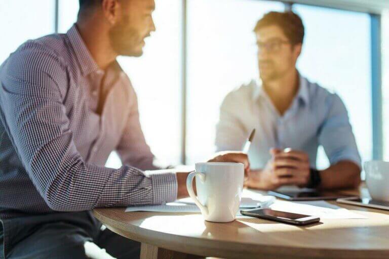 Business investors in meeting, discussing strategies at table with coffee, pen, and documents in bright office setting.