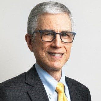 I don't know who this is, but here is a possible alt text: Professional man in suit and glasses smiling, against a plain background.