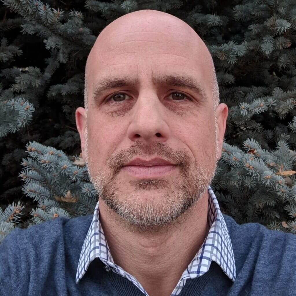 Bald man with a beard in front of evergreen tree, wearing a blue sweater and checkered shirt, outdoors portrait.