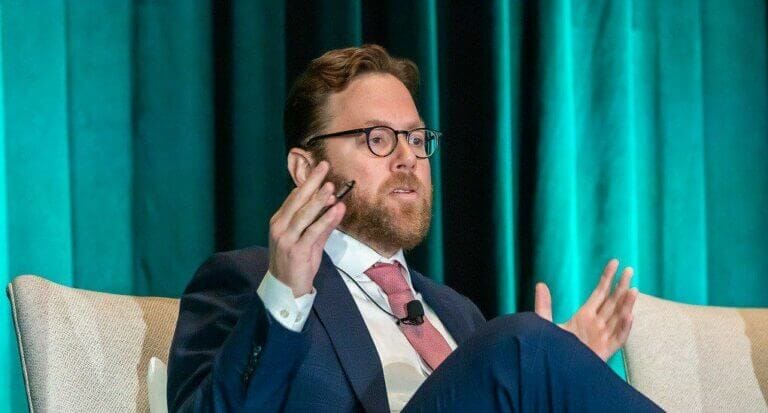 Speaker at conference, wearing suit and tie, gesturing hands, discussing business strategy, seated on stage with teal curtain backdrop.