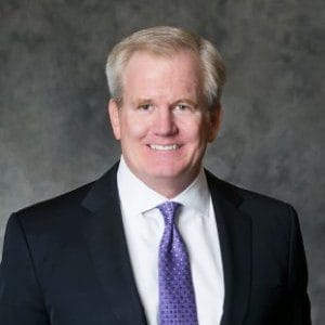 Professional man in suit with purple tie, smiling against a gray background. Business portrait, formal attire, corporate setting.