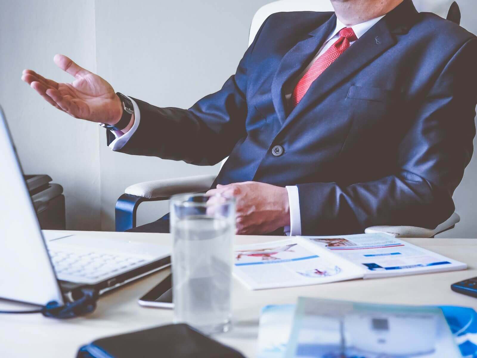 Businessman in suit gesturing during interview, sitting at desk with laptop and documents, professional meeting setting.