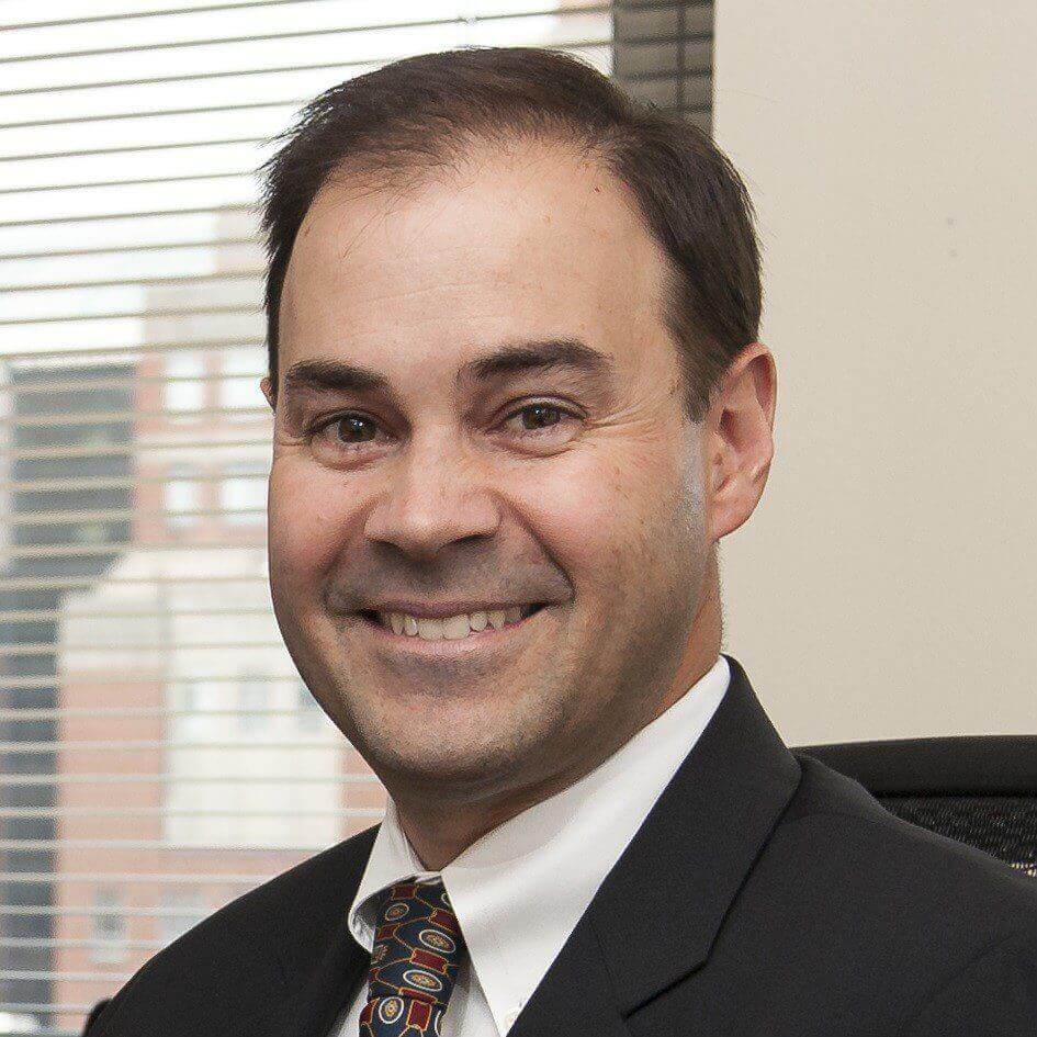 Smiling man in suit and tie in office setting with city view through window blinds.