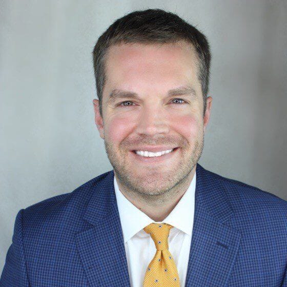 Smiling man in blue suit and yellow tie against gray background, professional portrait.