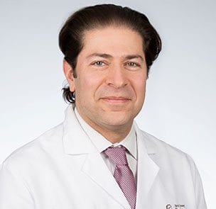 Professional man in a white lab coat and tie, smiling, against a neutral background, medical expert portrait.