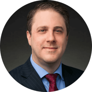 Portrait of a man in a suit with a red tie, against a dark background, professional headshot for business profile.