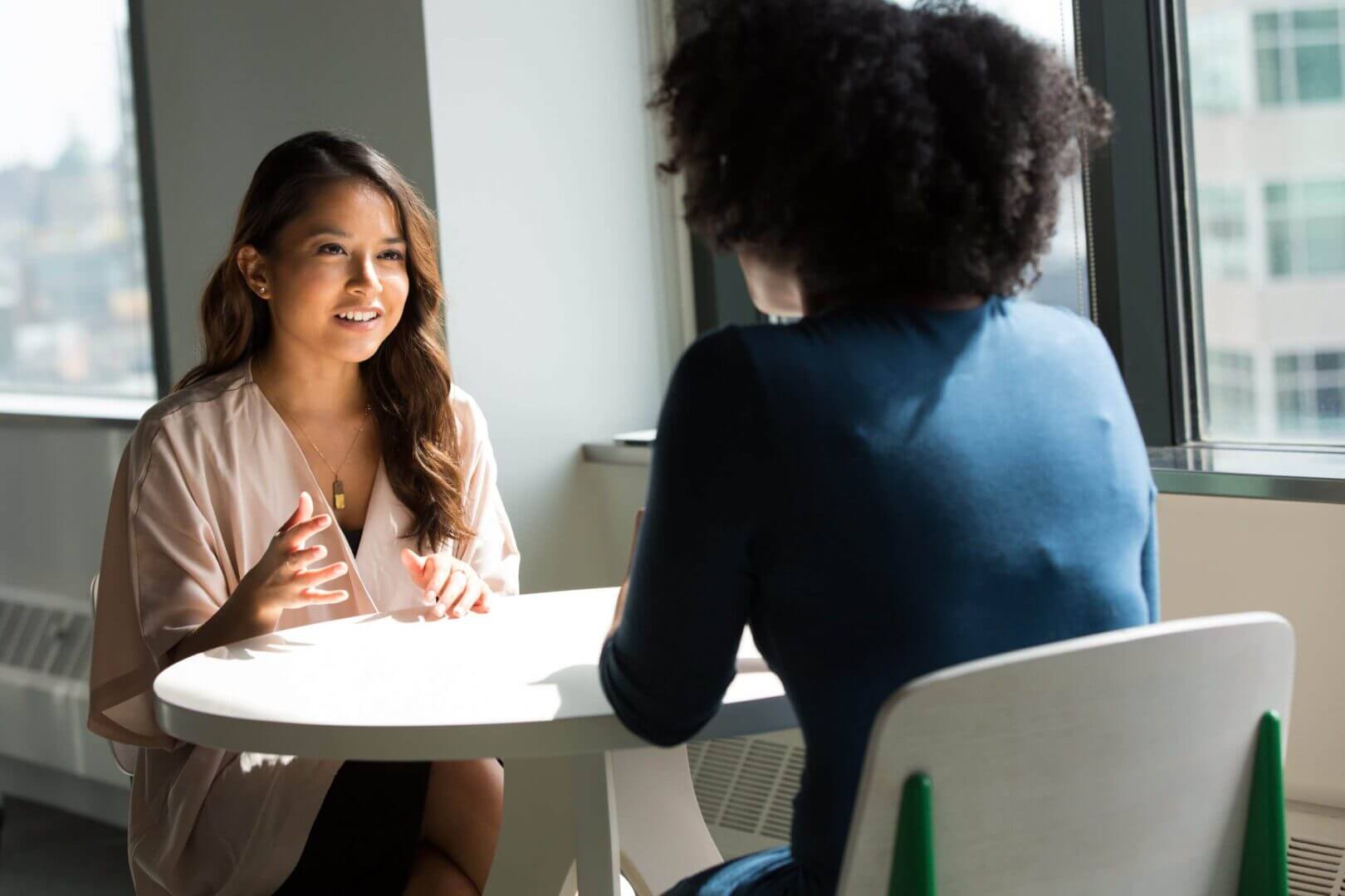 Two women having a business meeting at a round table, discussing professional tips in a bright office setting.