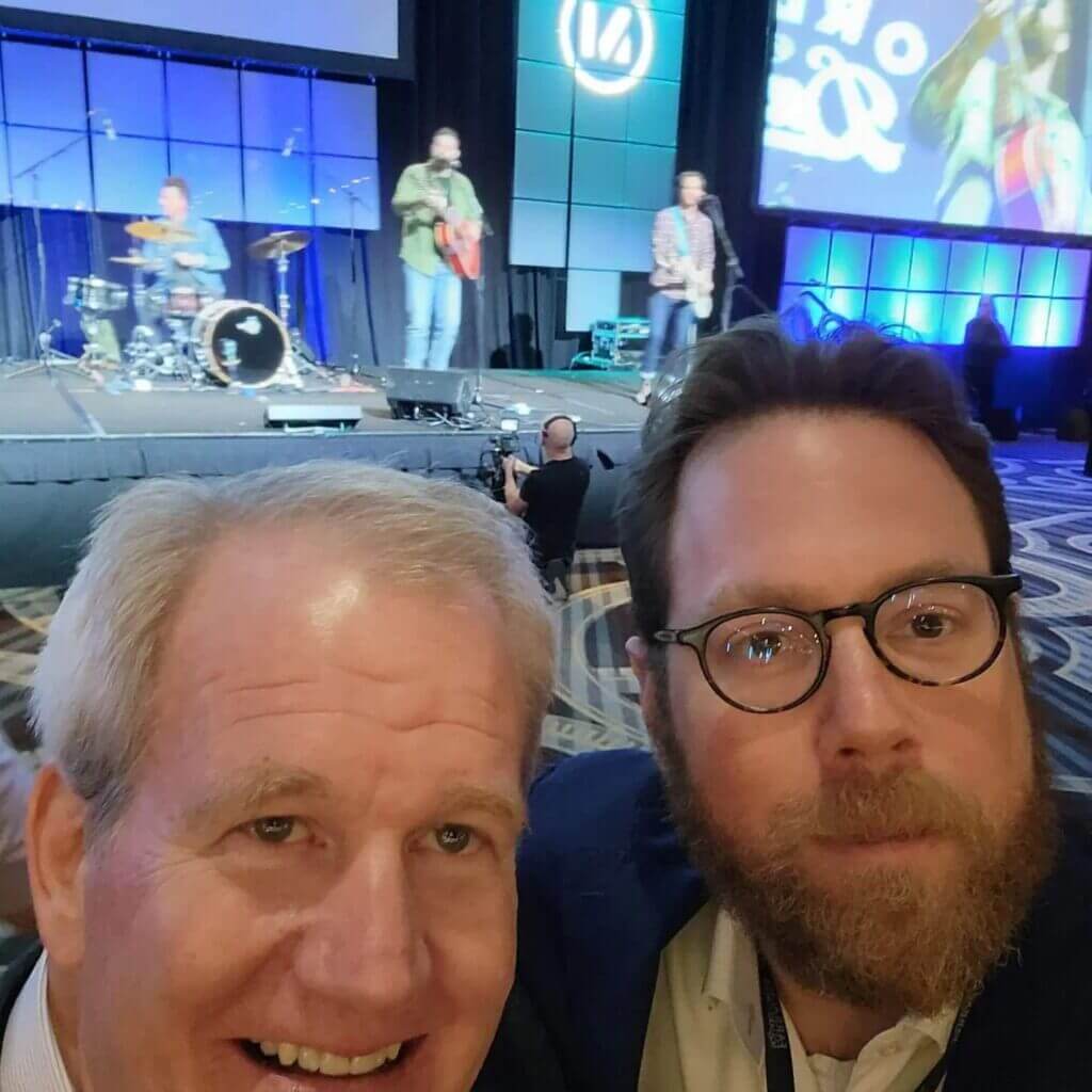 Two men taking a selfie at a live music event with a band performing on stage in the background.