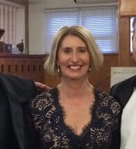 Middle-aged woman smiling in elegant lace dress at indoor event, standing between two people, soft lighting, wooden background.