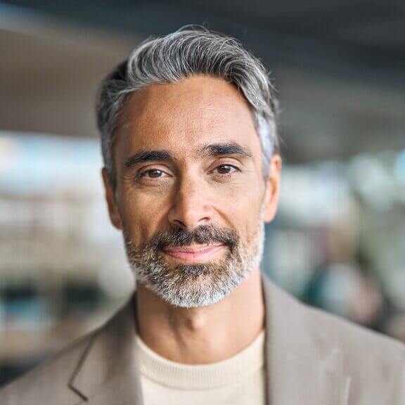 Smiling mature man with gray hair and beard wearing a beige blazer, outdoor professional portrait.